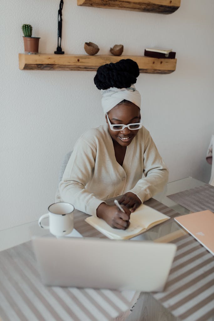 African American woman sitting indoors writing in a journal at a desk with a laptop and coffee mug.